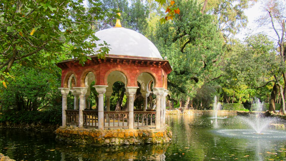 A decorative gazebo with a white dome and red-brick base stands in a lush, green park with fountains and trees.