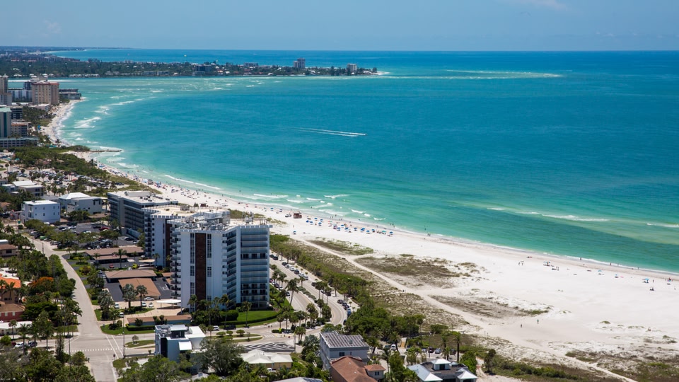 Aerial view of a coastal city with a long sandy beach and clear blue ocean, flanked by modern buildings and residential areas.