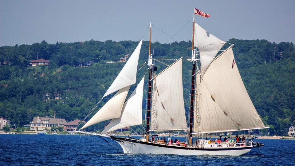 A large white sailboat with multiple white sails and a red flag on the mast, sailing on a blue lake with a forest-covered hill in the background.