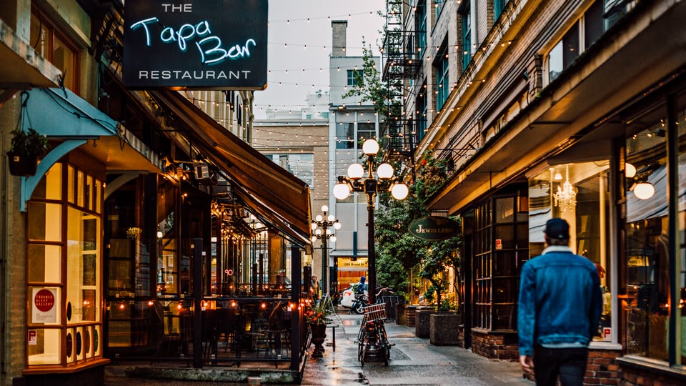 A narrow city street at dusk with a bar and restaurant signage, glowing string lights, and a person walking away.