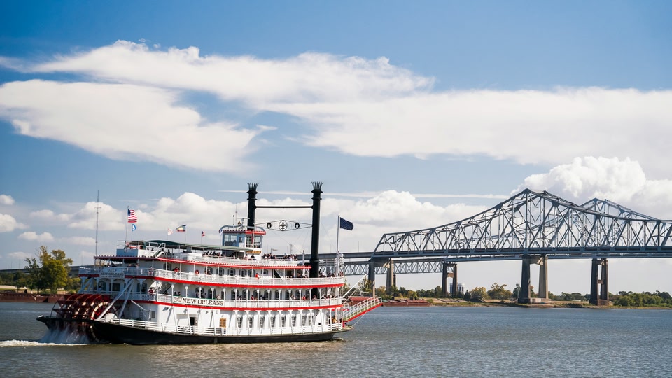 A large paddle steamer named City of New Orleans cruises along the river with a historic bridge in the background.