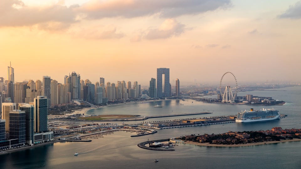 Aerial view of The Palm Jumeirah in Dubai at sunset, showing a skyline of modern buildings, a large cruise ship in the water, and the iconic Ain Dubai Ferris wheel.