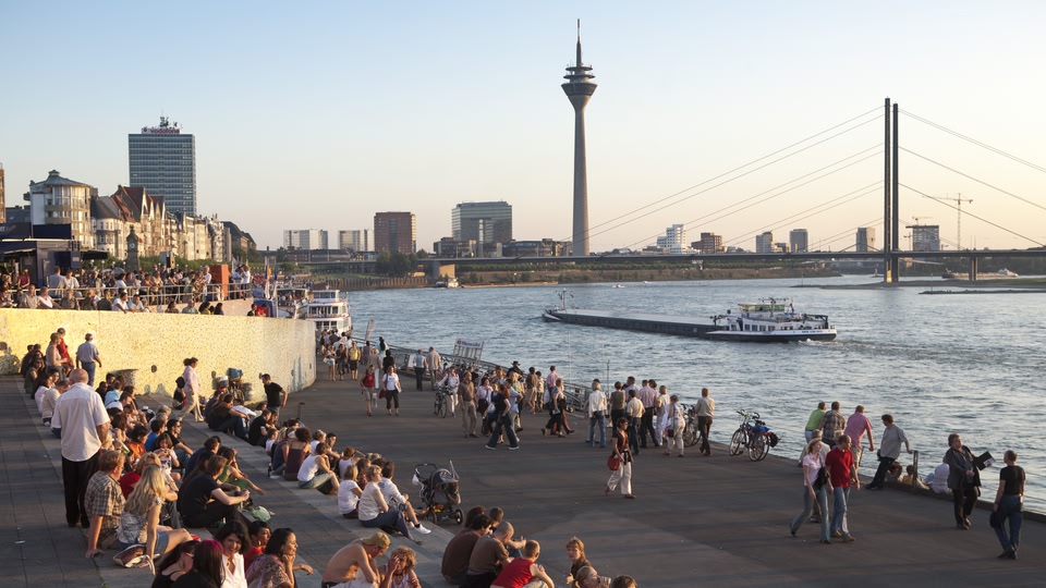 Crowd of people on a riverside promenade with boats in the water during sunset in Dusseldorf.