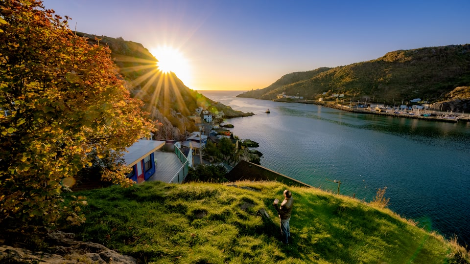 A person stands on a grassy hill overlooking a serene coastal town at sunrise, with the sun shining brightly in the background.