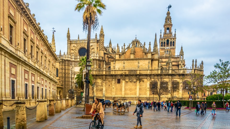 A historic plaza with ornate buildings and people walking and cycling around.