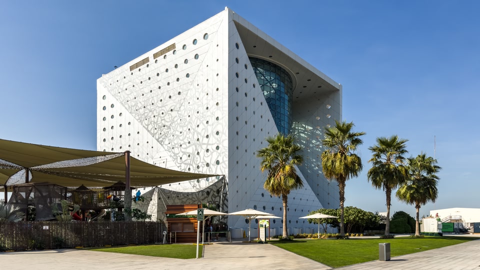 A modern, uniquely designed white building with circular windows and a curved glass section, set against a clear blue sky. The building is surrounded by palm trees and green landscaping.