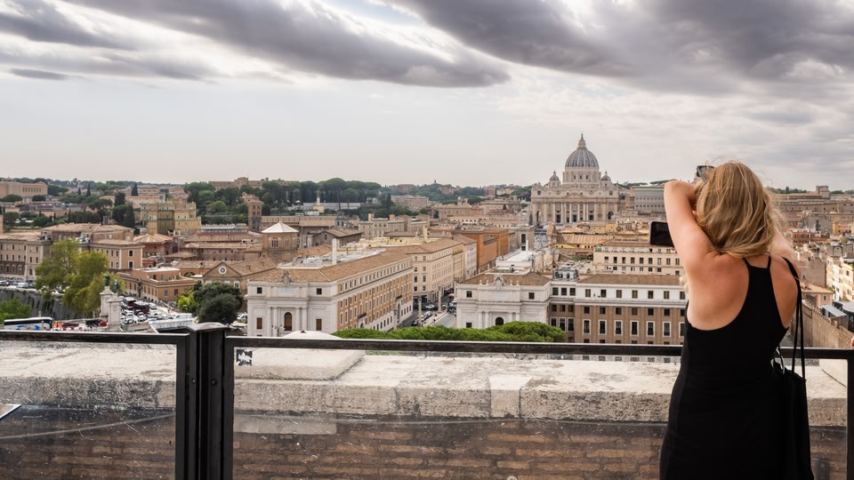 A woman stands on a balcony capturing a panoramic view of Rome, Italy, with St. Peter's Basilica prominently visible in the distance.