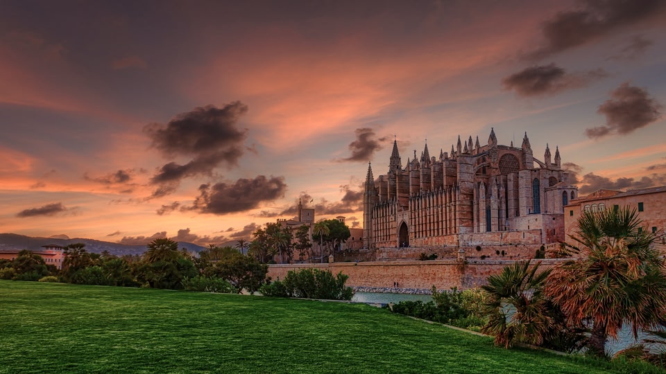 A large cathedral with multiple spires, set against a dramatic sunset with a green park in the foreground.