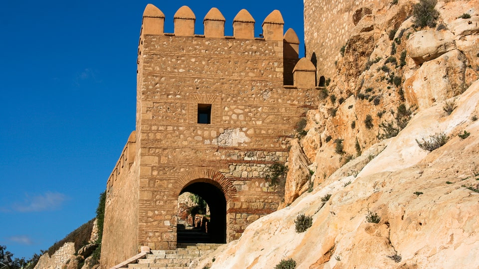 An ancient stone archway and tower built into a rocky hillside under a clear blue sky.
