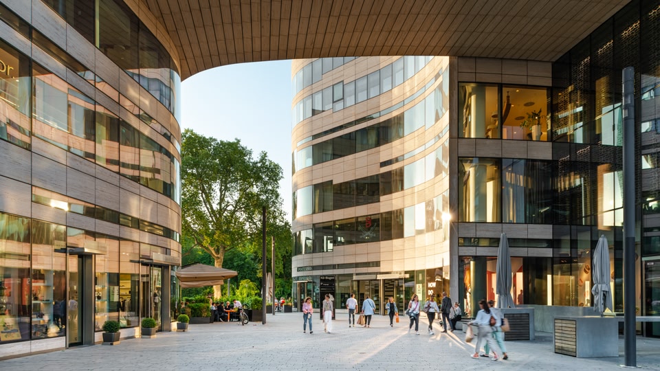A bustling urban shopping street with modern buildings, people walking, and storefronts visible.