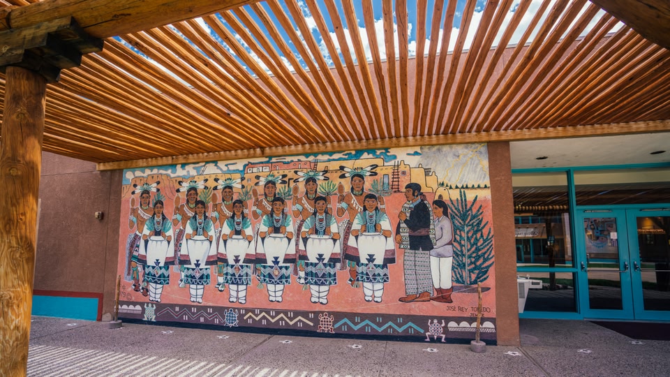 A mural depicting traditional Native American dancers and scenes is painted on the side of a building with a wooden canopy in Albuquerque, New Mexico.