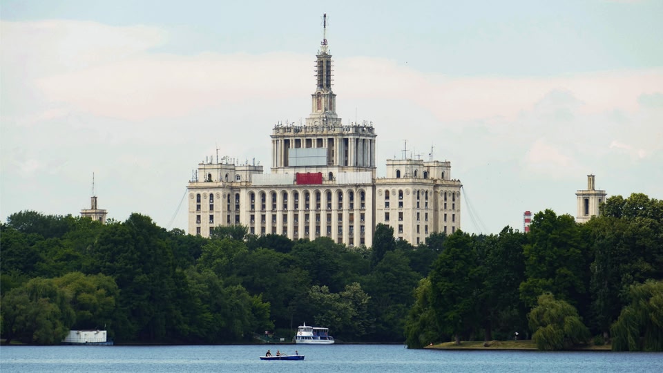 A large, imposing building with a central tower surrounded by trees and overlooking a lake.