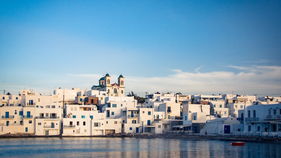 A view of the picturesque white buildings of Naoussa town in Greece, with a church in the background and the sea in the foreground.