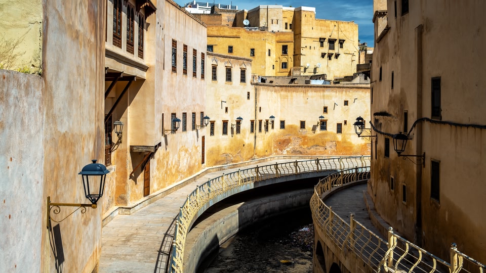 Curved walkway and iron lamps along ochre plaster buildings over a narrow stream in Fes.