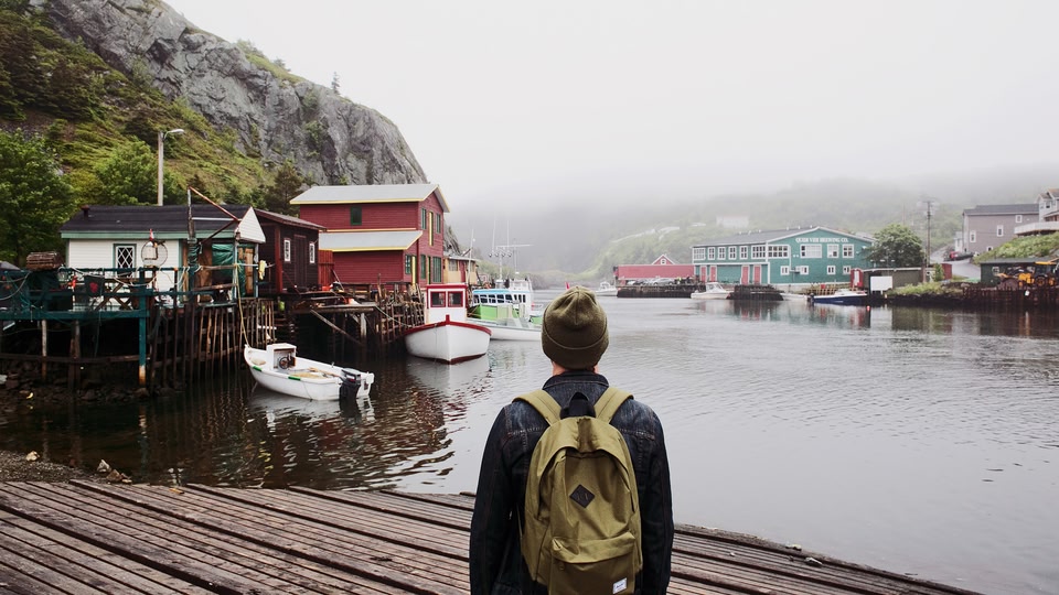 A solo traveler with a backpack stands at the edge of a wooden dock, gazing out at a harbor with colorful houses and moored boats.