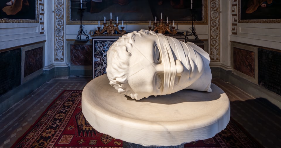 A white sculpture of a head on a pedestal inside a museum room with ornate decor.