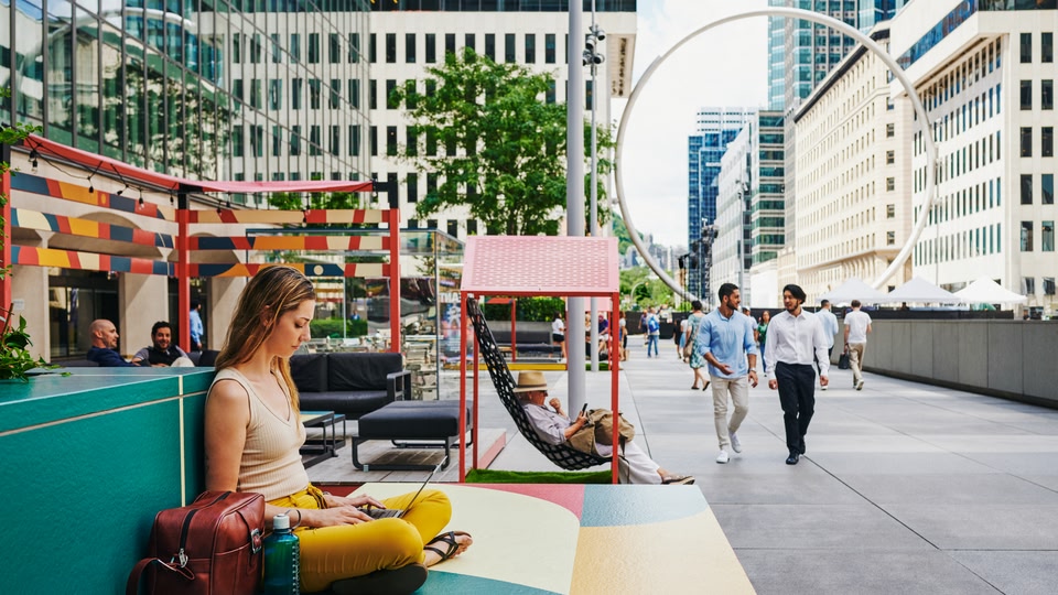 A woman sitting on a colorful outdoor bench using a laptop, with several people walking and socializing in an urban setting.