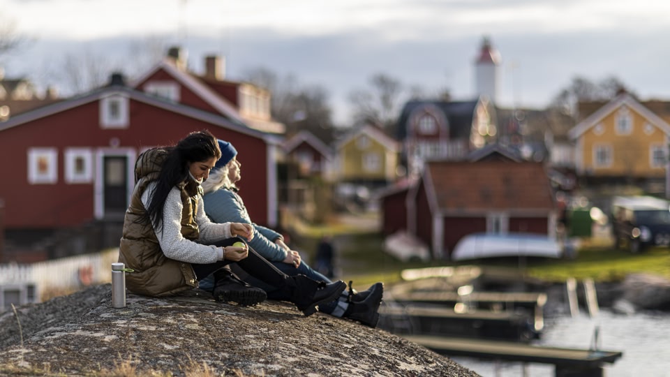 Two people sitting on a rock overlooking a village with colorful houses and a calm water body in the background during sunset