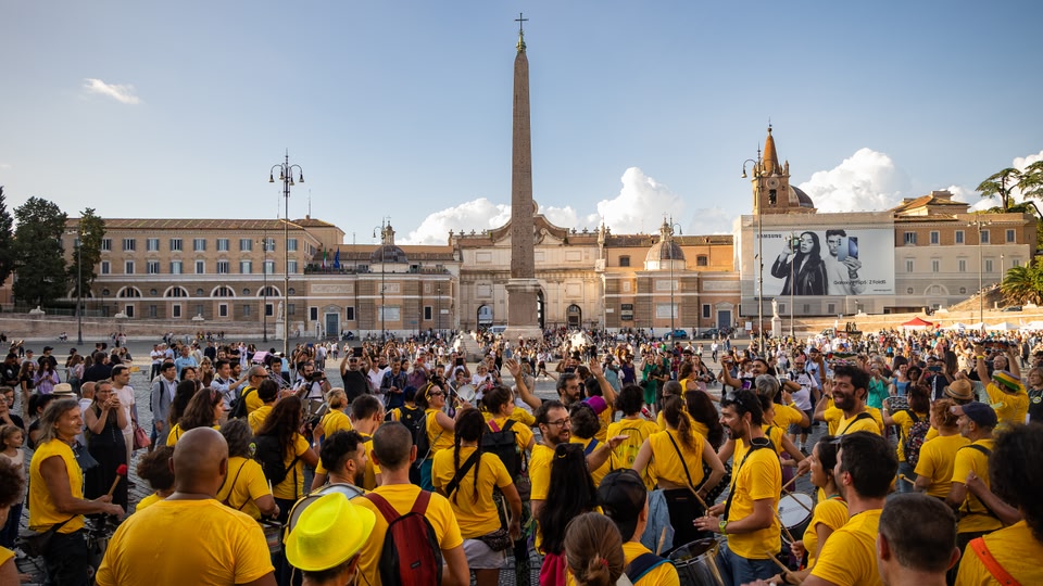 A large crowd of people in yellow shirts gathered in a city square with musical instruments, in front of historical buildings under a clear blue sky.