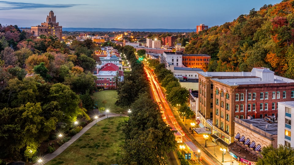 Aerial view of a city at dusk with autumn-colored trees and a long stretch of illuminated road leading into the city center.