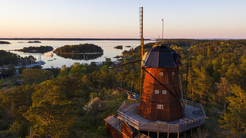 Aerial view of an old windmill with a wooden platform and flagpole, surrounded by lush greenery and overlooking a serene body of water at sunset.