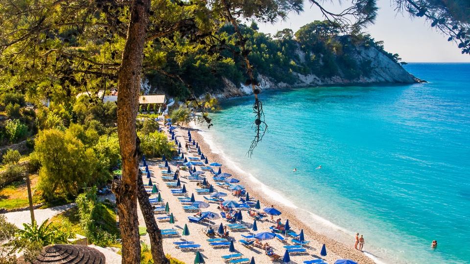 A sunny beach scene with rows of blue sun loungers and umbrellas set up on the sand, overlooking a clear blue sea.