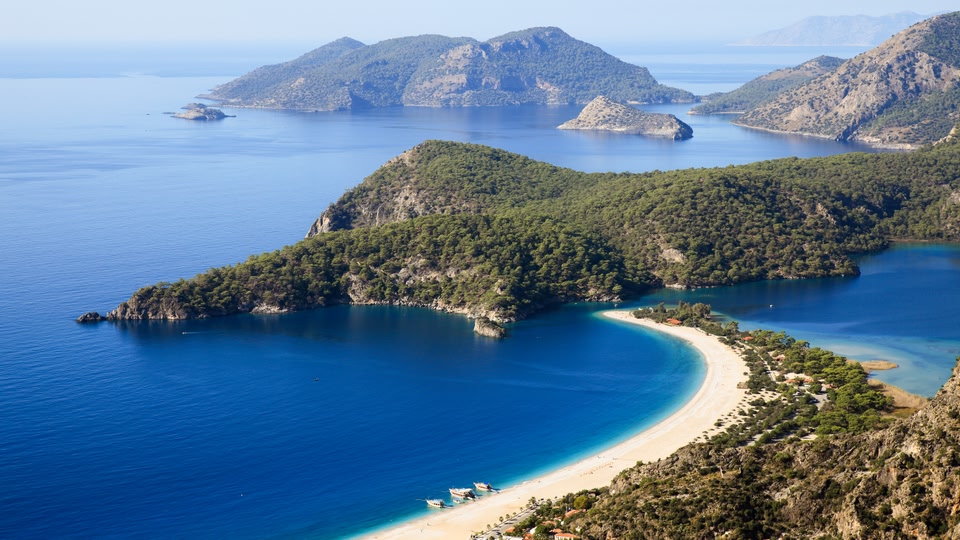 Aerial view of a turquoise bay with a sandy beach and forested hills in Dalaman, Türkiye.
