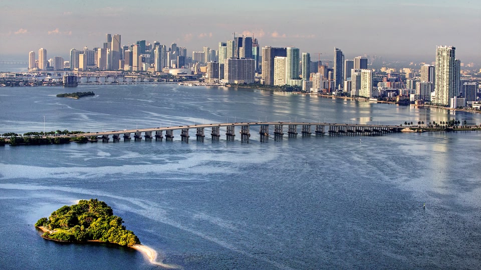 Aerial view of Miami, Florida, showcasing the city skyline and a bridge over water with a small island in the foreground.