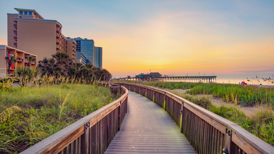 A wooden boardwalk leading through grassy dunes towards a beach and pier at sunset.