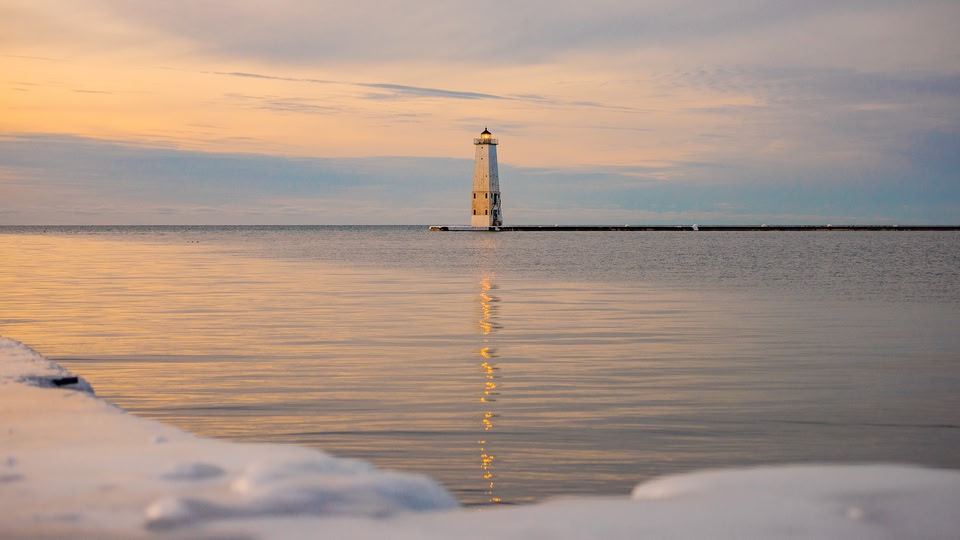 A lighthouse standing in the middle of a body of water with a snowy foreground, captured during sunset.