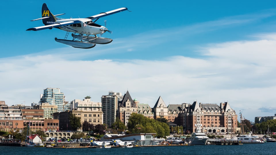 A seaplane flying above the skyline of Victoria, British Columbia, with various boats docked below.