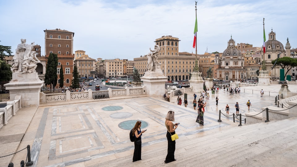 A bustling open square with historical buildings, monuments, and many people walking around.