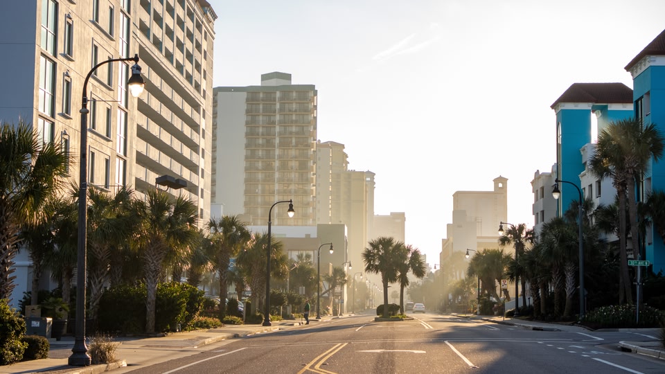 A city street at sunrise or sunset with tall buildings, palm trees, and street lights