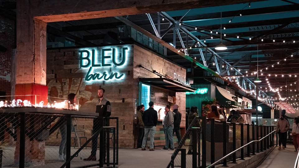 Outdoor seating area of Bleu Barn with people standing and a large sign lit up above.