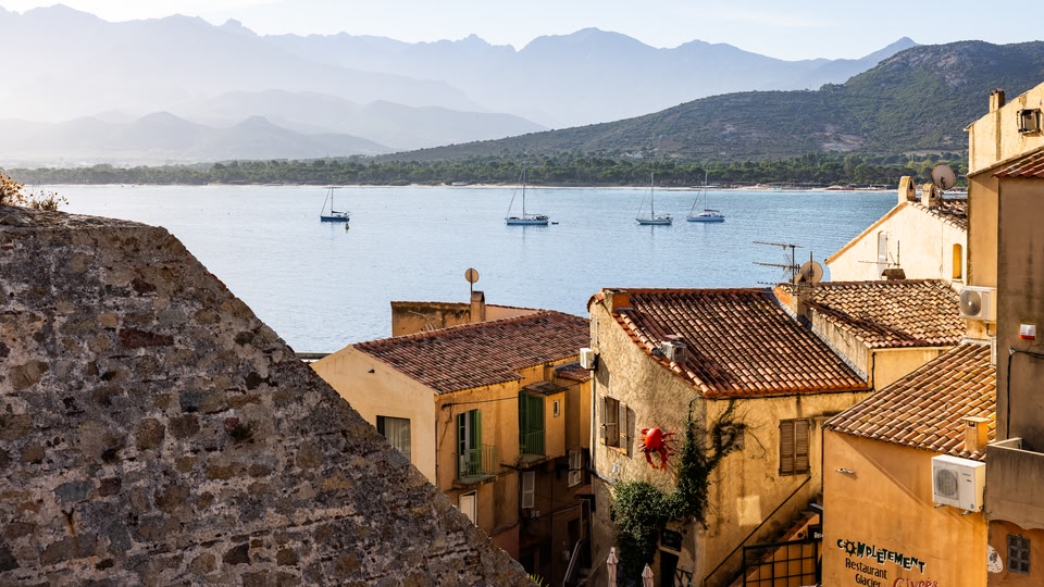 Rooftops with terracotta tiles and a red crab sculpture above a calm bay with sailboats in Calvi.