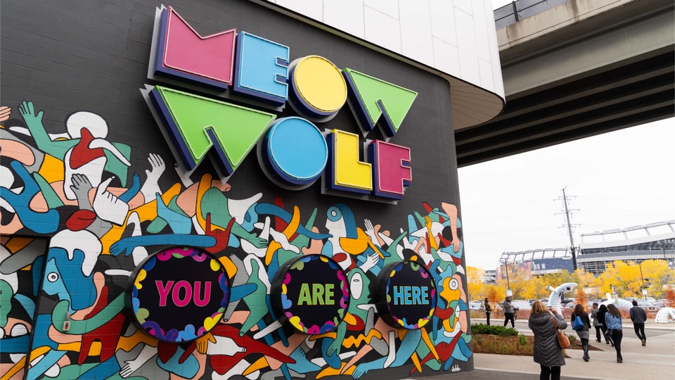 A colorful mural titled 'MEOW WOLF' on the side of a building in an urban setting with people walking by.