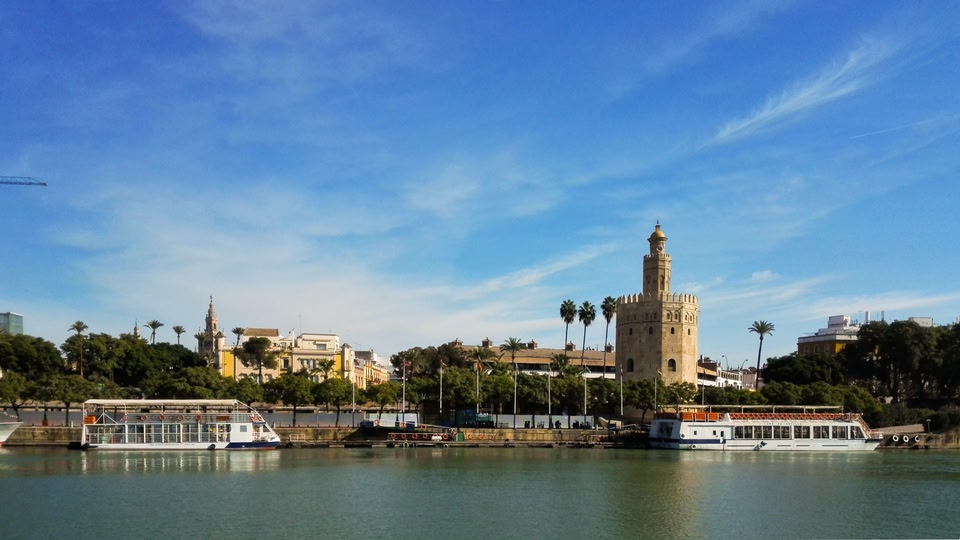 A scenic view of Seville, Spain, with boats on the river and the iconic Torre del Oro tower in the background.
