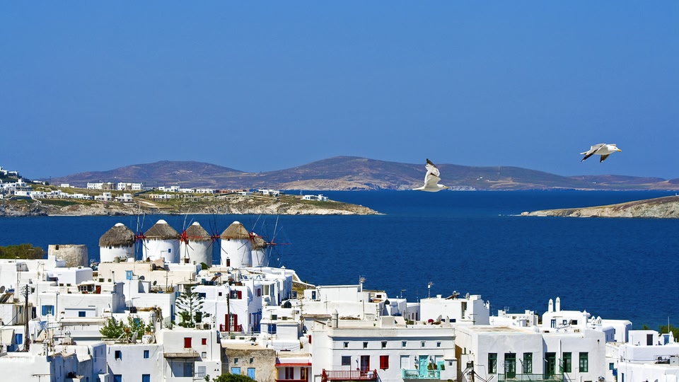 Aerial view of a coastal town with white buildings and windmills, overlooking the blue sea.