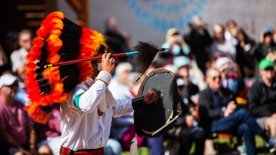A performer in a feathered headdress and holding a drum, engages with the audience at an outdoor event.