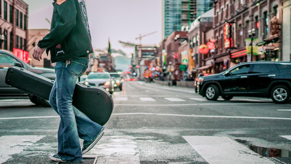 A person carrying a guitar case walks across a city street with vibrant neon signs in the background.