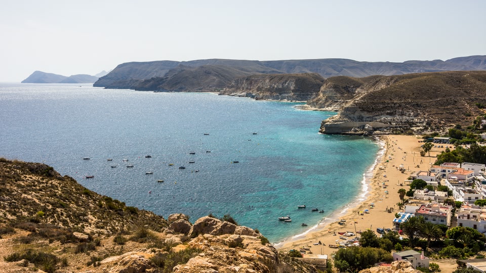 Aerial view of a coastal beach with clear blue waters, boats, and a nearby town in Almeria, Andalusia, Spain.