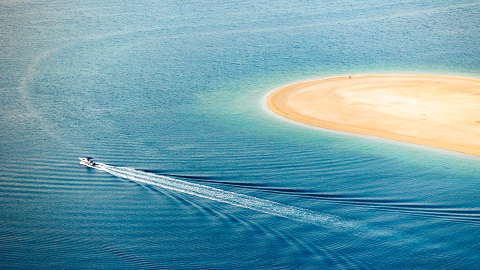 Aerial view of a boat sailing near a small sandy island in clear blue waters.