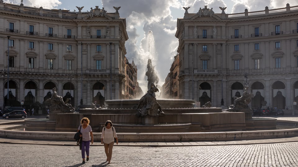 Two women walking past a historic fountain in Rome, Italy.