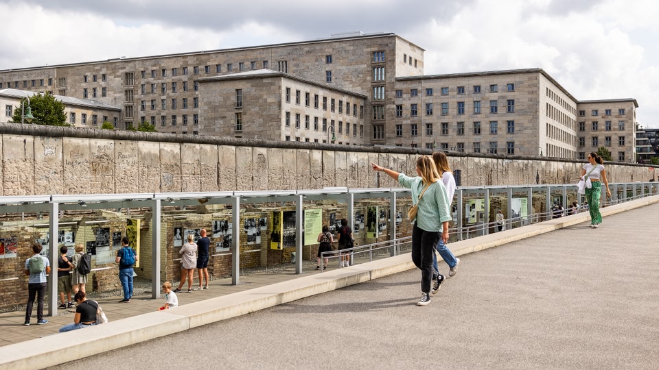 Visitors exploring the outdoor exhibits at the Topography of Terror museum in Berlin, with a large, graffiti-covered wall and a modern building in the background.