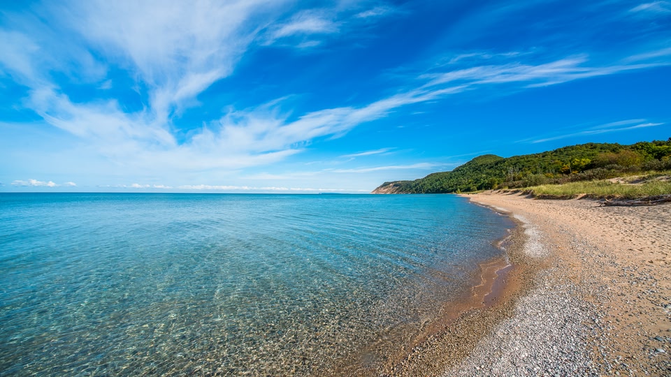 A serene beach scene with clear blue waters and a rocky shore under a partly cloudy sky.
