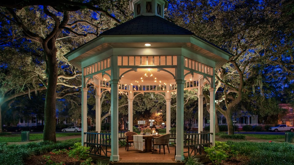 A gazebo with a table set for a night-time dinner, surrounded by trees and illuminated by fairy lights in a park.