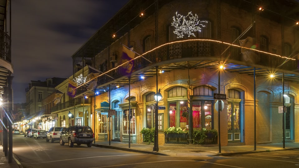 Nighttime view of a historic street in New Orleans with illuminated buildings, decorative lights, and parked cars.