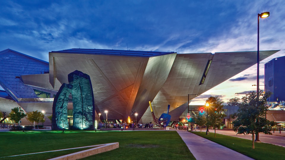 The Denver Art Museum at sunset with blue sky and illuminated facade, surrounded by green lawns and trees.