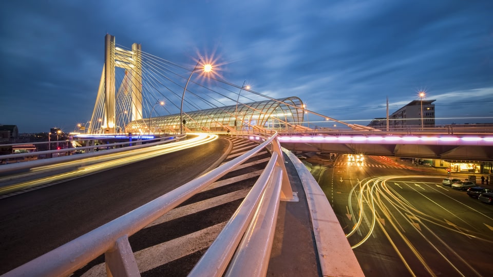 A nighttime view of a bridge and cityscape with glowing lights and blurred car trails.