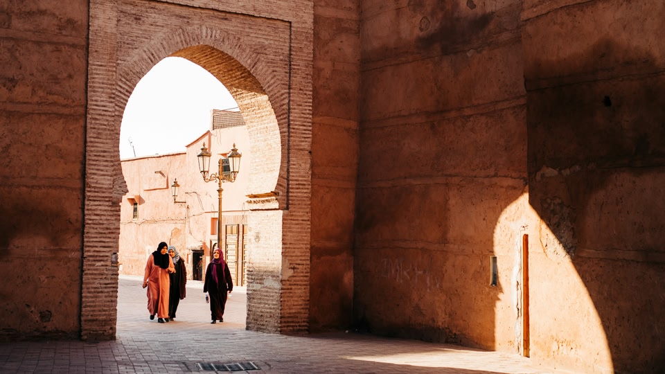 Three people wearing headscarves walk beneath a brick archway in Fes.